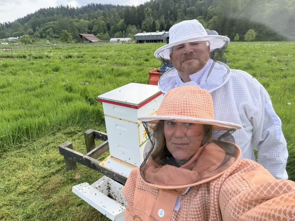 Kappie and Joe in beekeeping suits next to one of the hives — Kappie in the orange veil in front, Joe in the white veil behind, with the pasture and a barn visible in the distance
